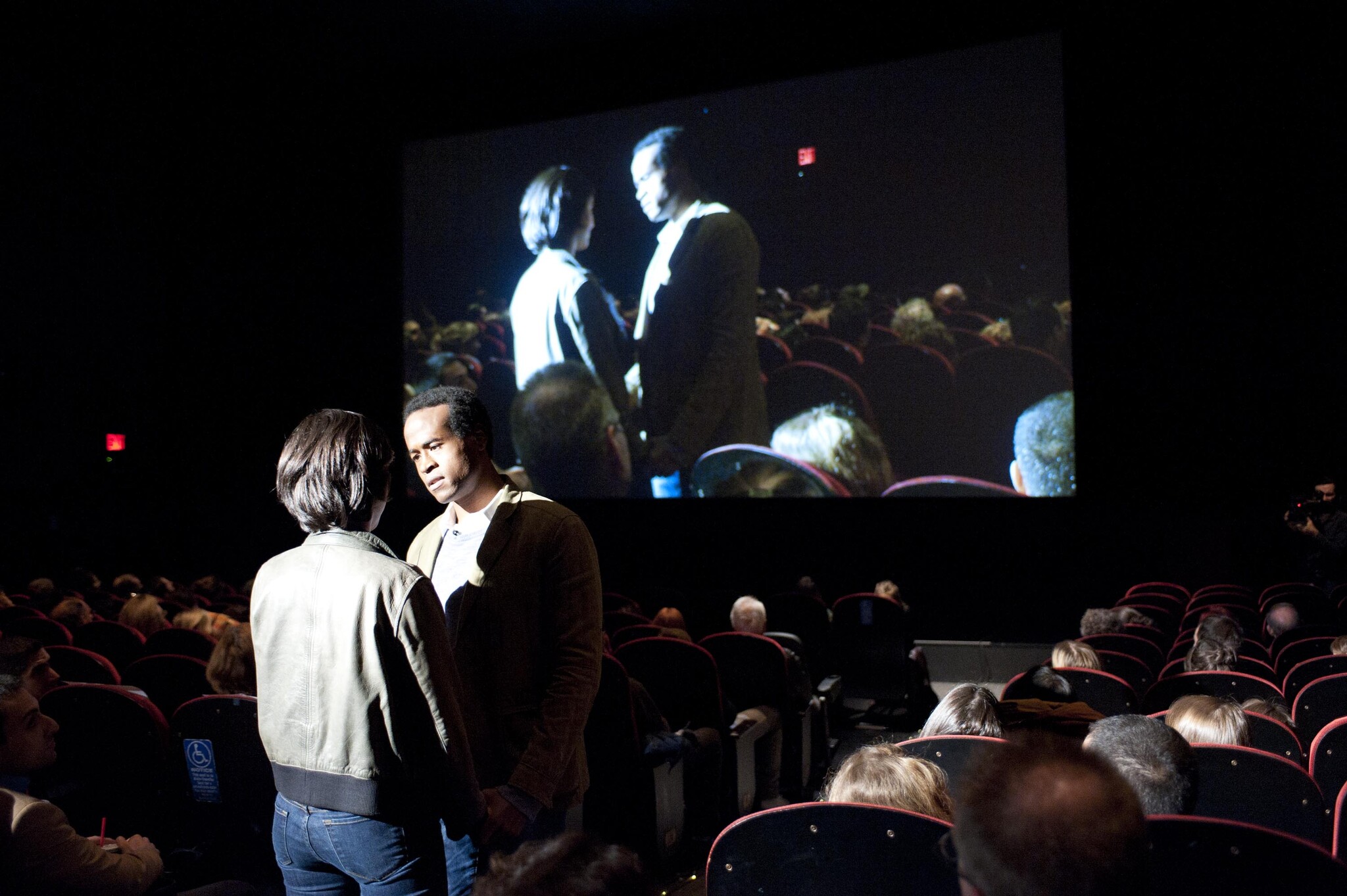 Two people stand facing each other in a dim movie theater aisle while the screen projects them.
