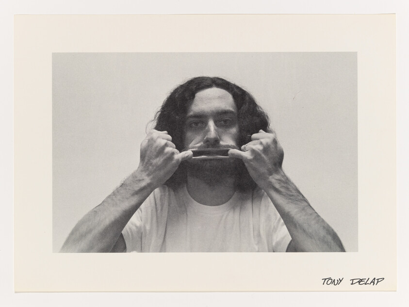 Man with shoulder-length hair stretches his mouth outward with both hands while facing the camera.