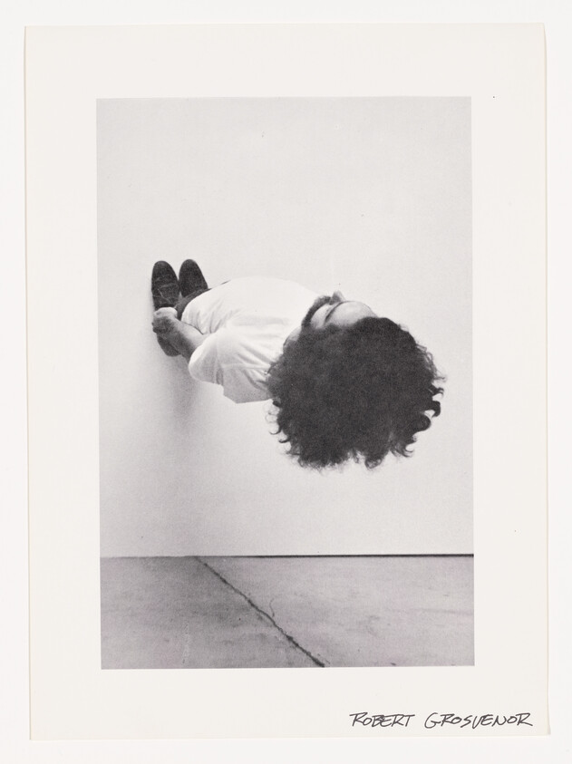 Man with curly hair suspended horizontally against a white wall, legs crossed and arms behind back.