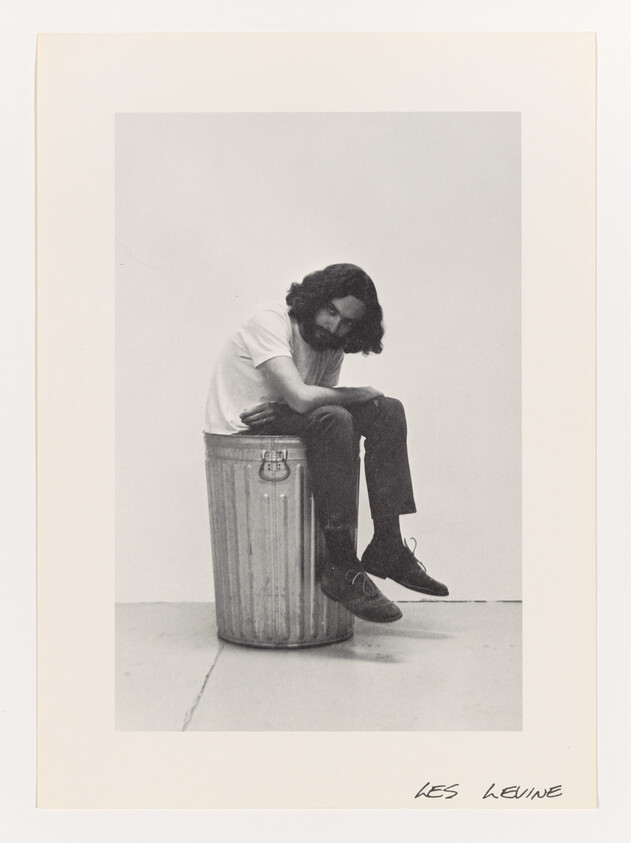 A man with long hair and beard sits hunched on a metal trash can.