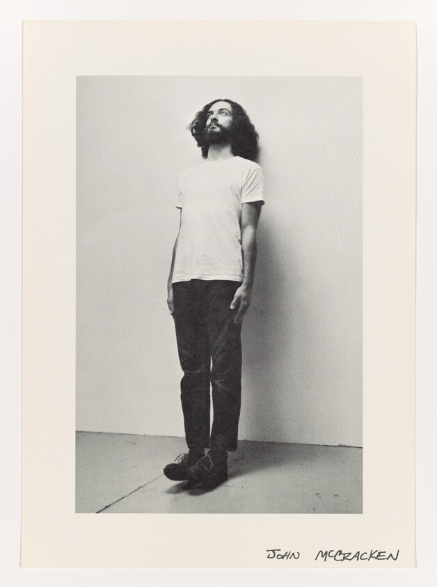 A bearded man in a white t-shirt and dark pants stands against a wall looking upward.