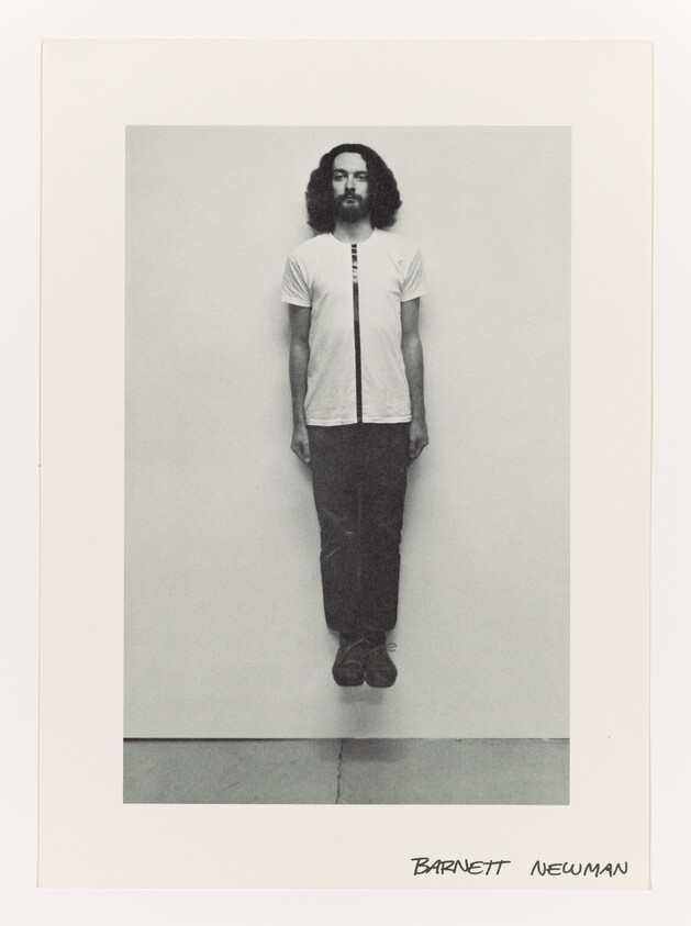 A man with long hair and a beard appears to float vertically against a plain wall.