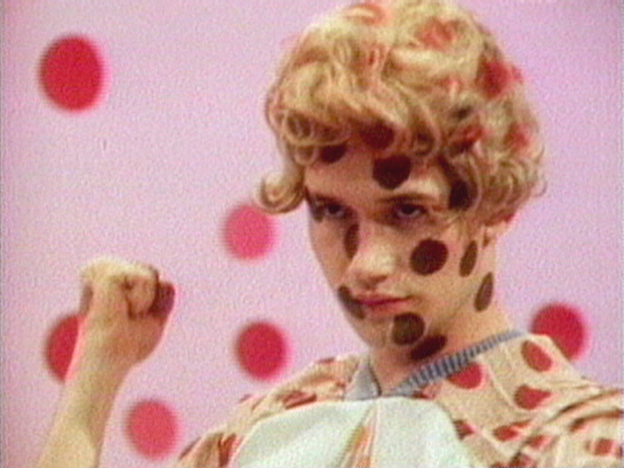 Young person with polka dots painted on face and clothing raises a fist against a dotted background.
