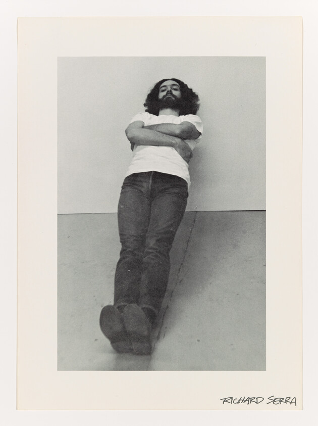 Bearded man leaning against a wall with arms crossed and legs extended toward the camera.