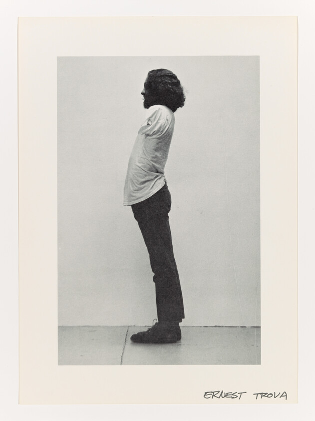 Man standing in side profile with hands behind his back against a plain background.