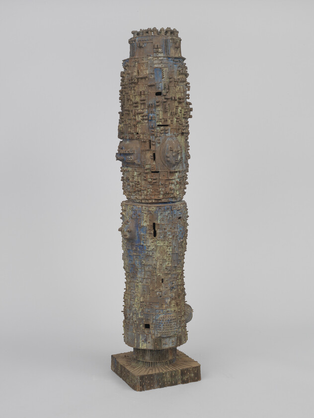 Tall weathered wooden sculpture with intricate carved geometric patterns standing on a square base.