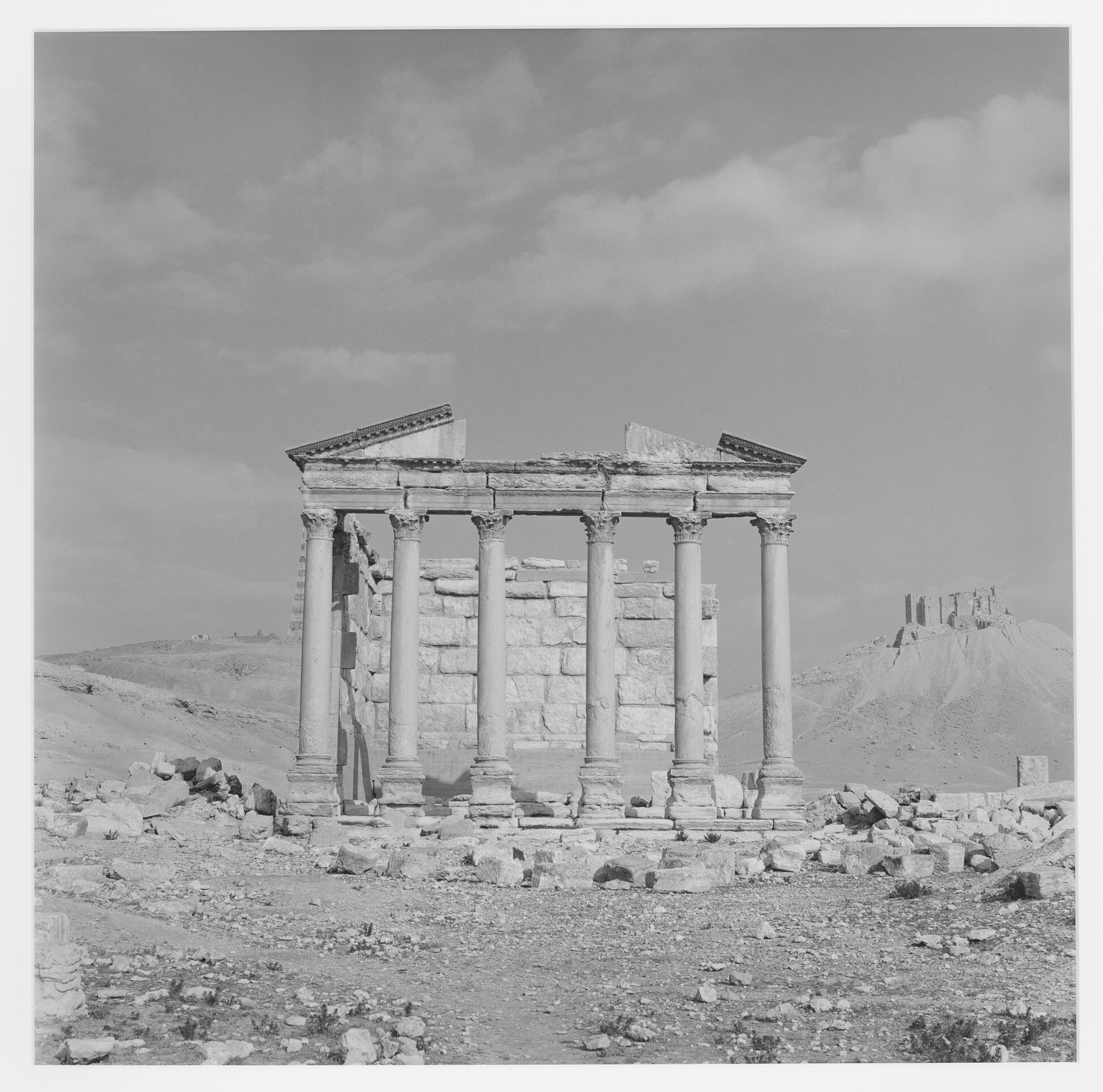 Six-columned ancient temple ruins standing in a rocky desert with a distant hilltop fortress.