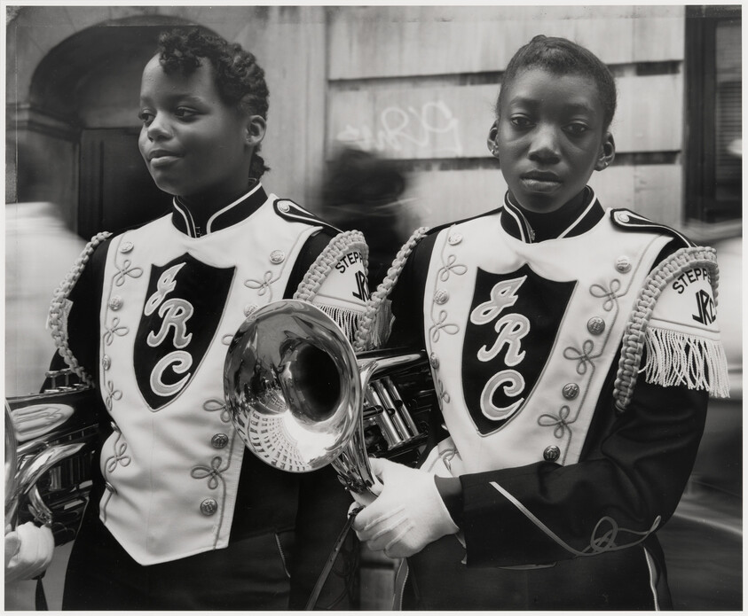 Two young marching band members in matching uniforms holding brass instruments on a city street.