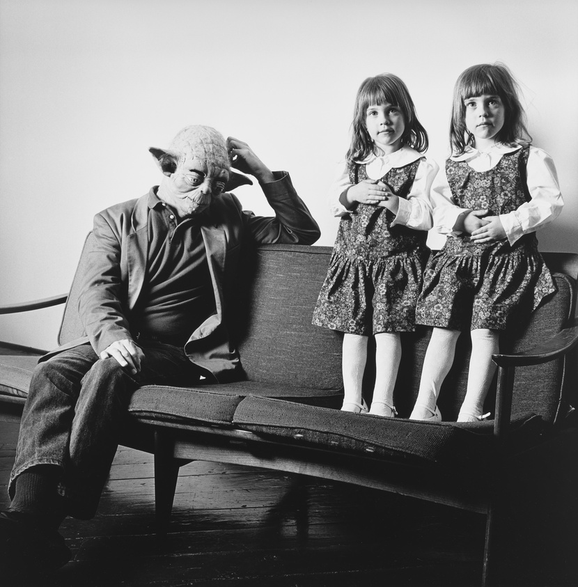 A black and white photograph featuring an individual wearing a Yoda mask sitting on a couch, scratching their head, flanked by two young girls in matching dresses and white tights, standing on the couch with their hands on their hearts.