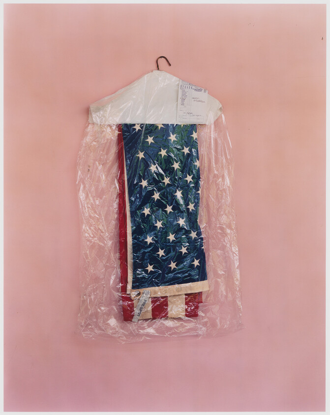 Folded American flag protected in dry-cleaning plastic hanging on a hanger against a pink wall.