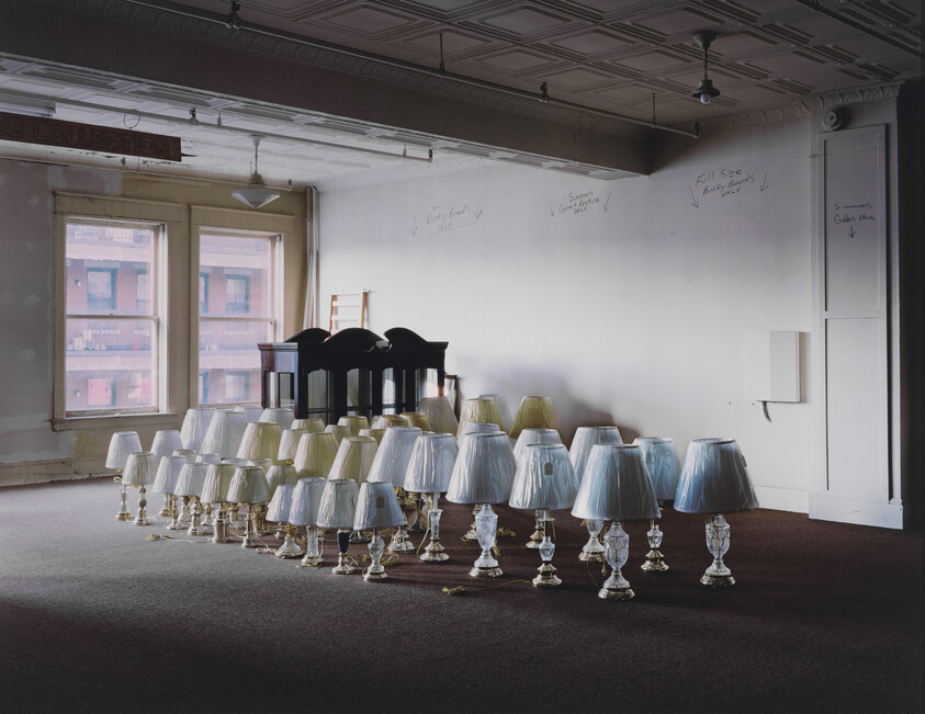 Dozens of glass table lamps with shades arranged in rows on a carpeted floor.