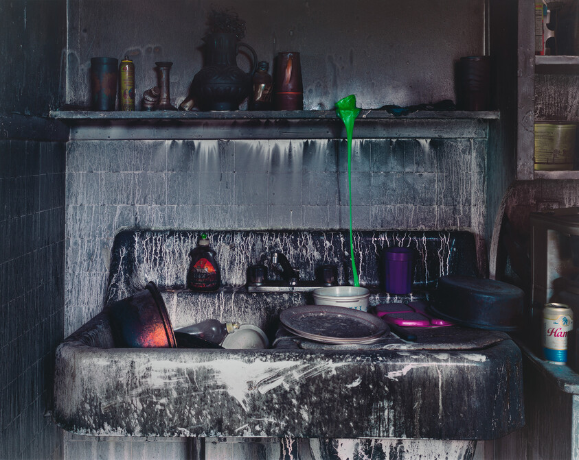 A grimy kitchen sink filled with dirty dishes while bright green liquid drips from a shelf above.
