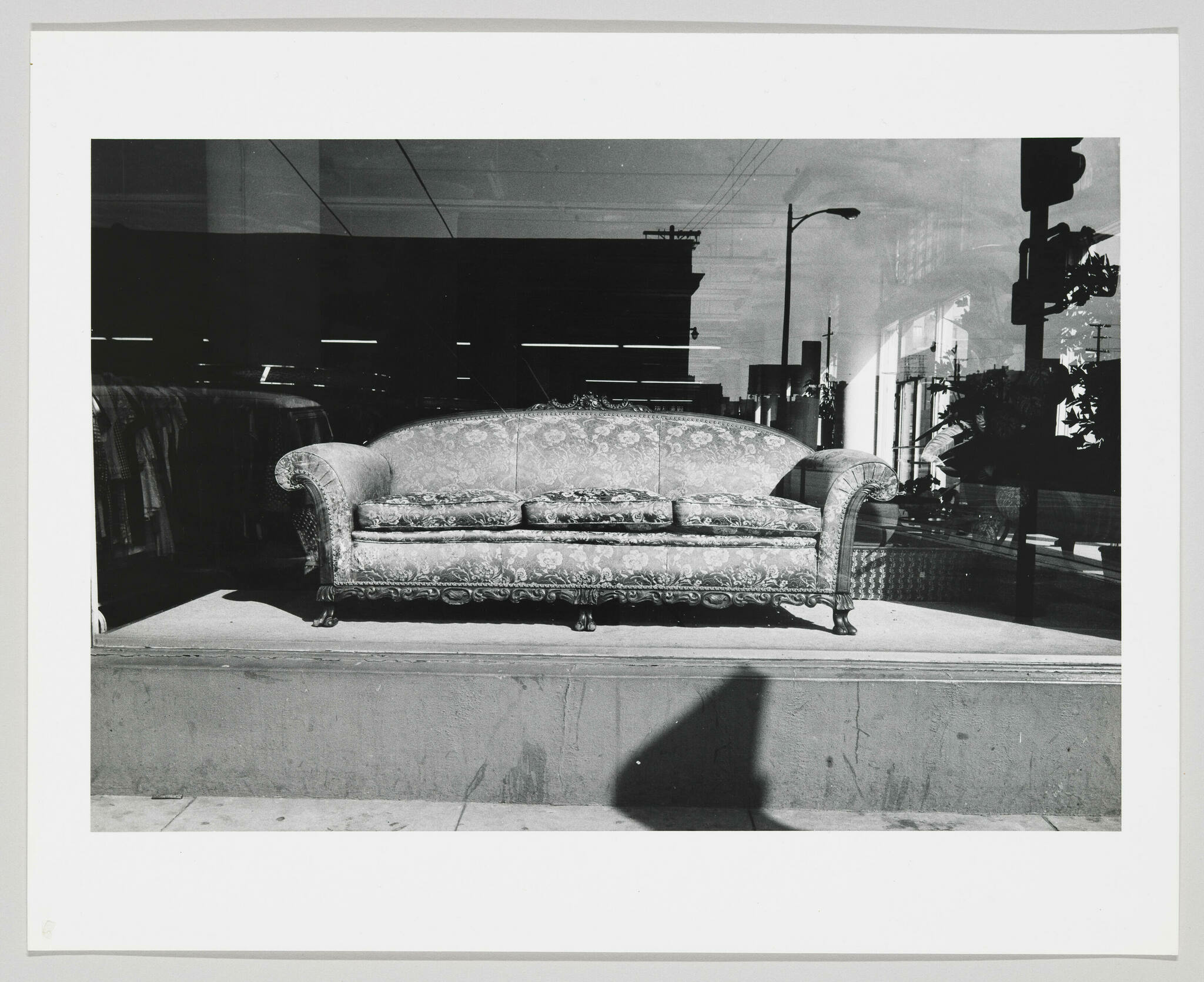 A black and white photograph showing an ornate couch displayed in a storefront window, with reflections of the street and a pedestrian's shadow visible on the glass.