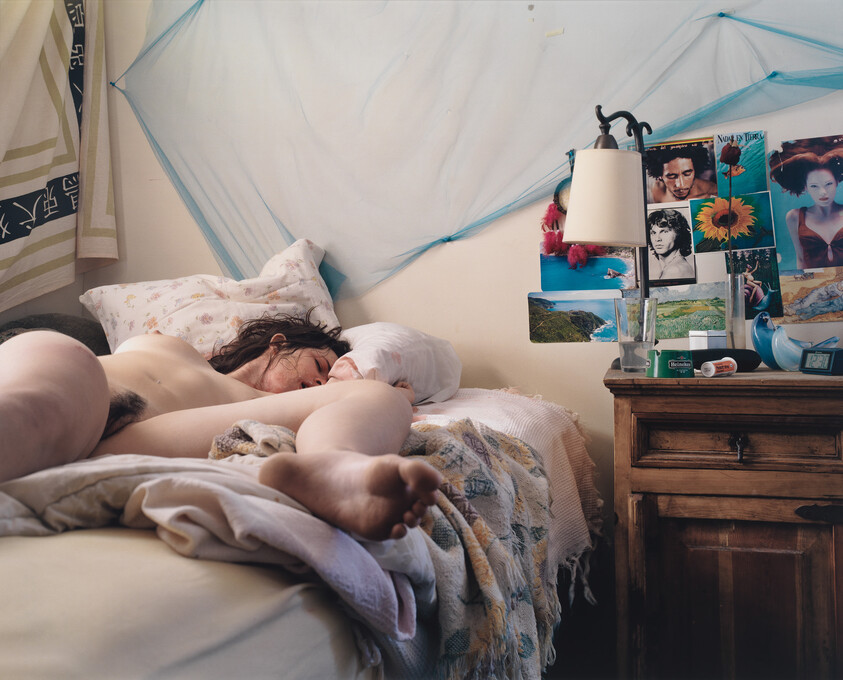 A person lies sleeping naked on a messy bed beside a wooden nightstand with photos.