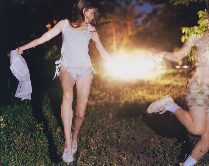 A young woman in a tank top and floral bikini bottoms runs through grass at night holding a shirt.