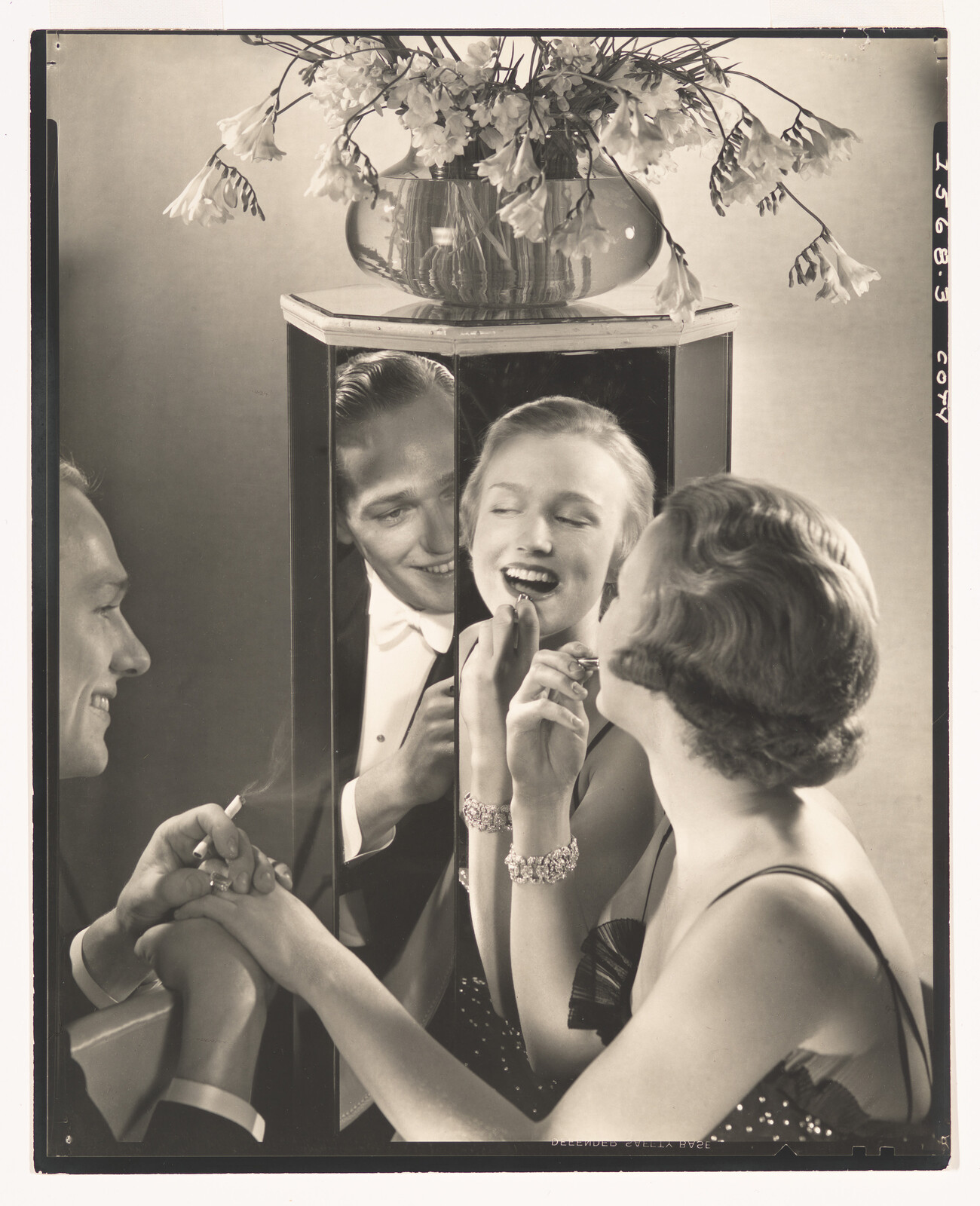 A woman applies lipstick while a smiling man holds her hand and watches through a mirror.