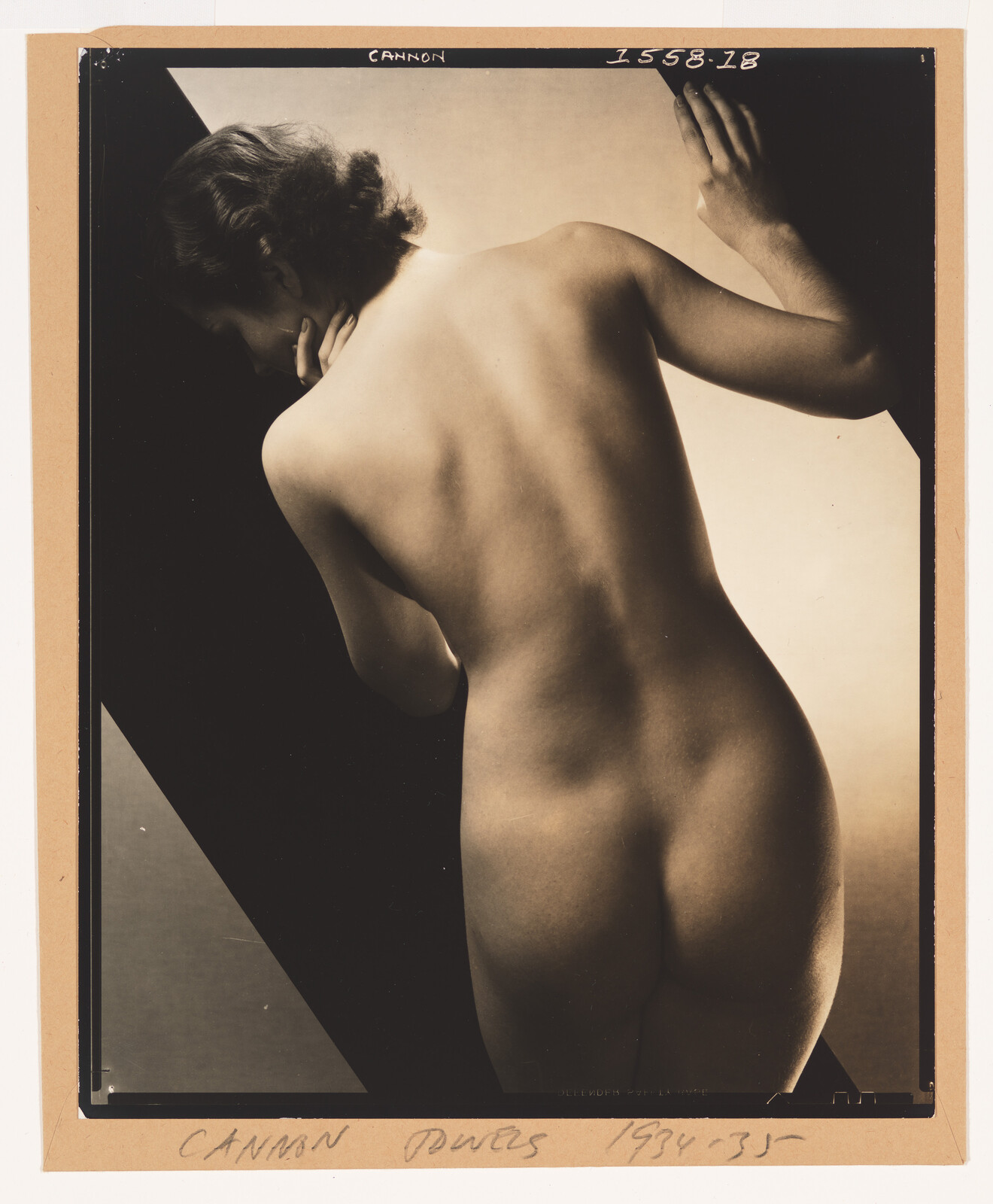 Nude woman seen from behind leaning against a dark panel, highlighting her back and hips.