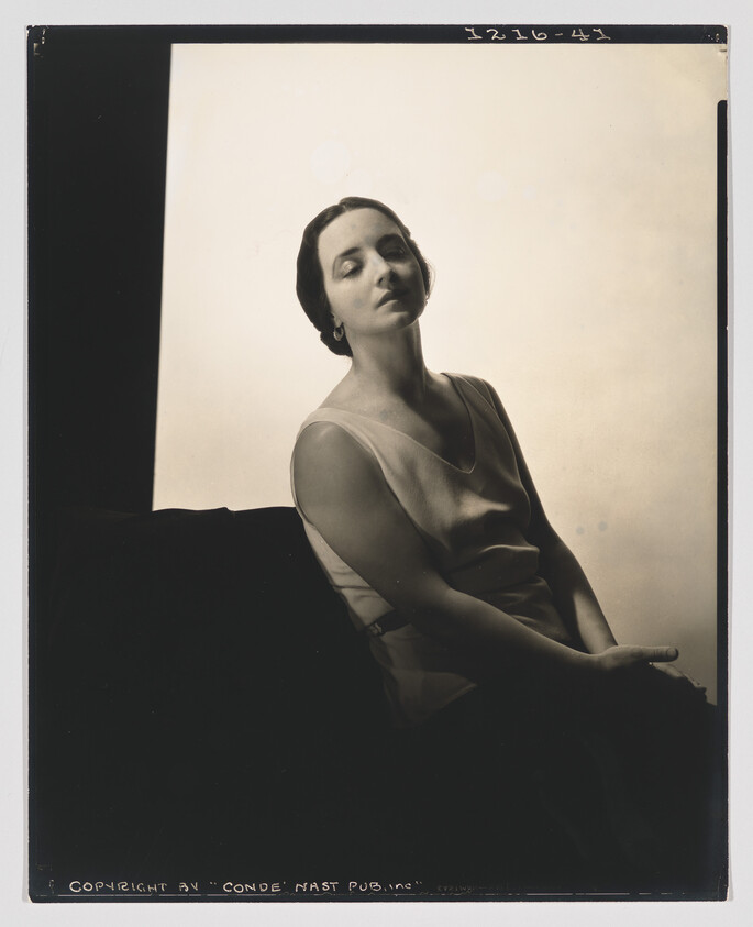 A seated woman in a sleeveless dress poses with closed eyes and relaxed, elegant posture.