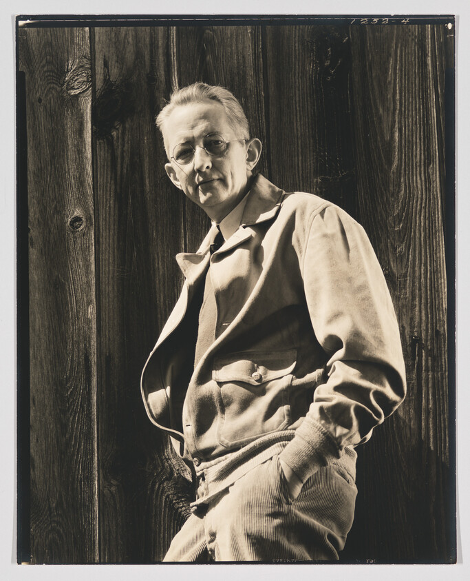 A man in glasses and a jacket stands leaning against a wooden wall with hands in pockets.