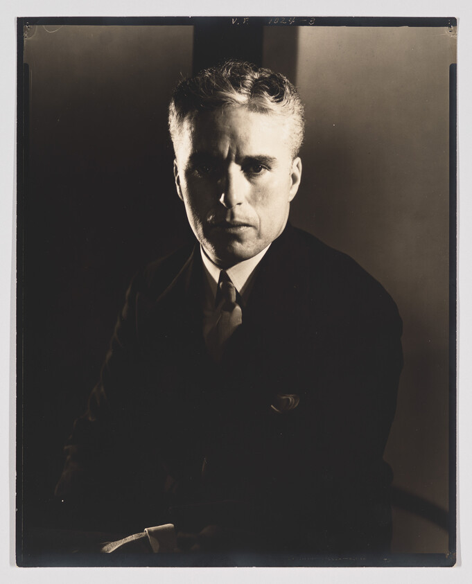 A serious man in a dark suit and tie stares directly at the camera in dramatic lighting.