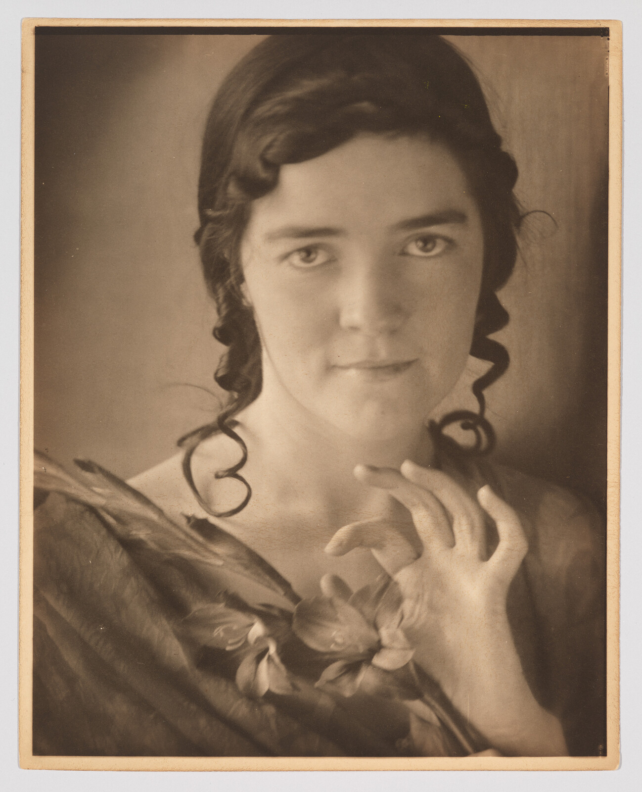 Young woman with braided ringlets holds a flower near her chest and gently gestures.