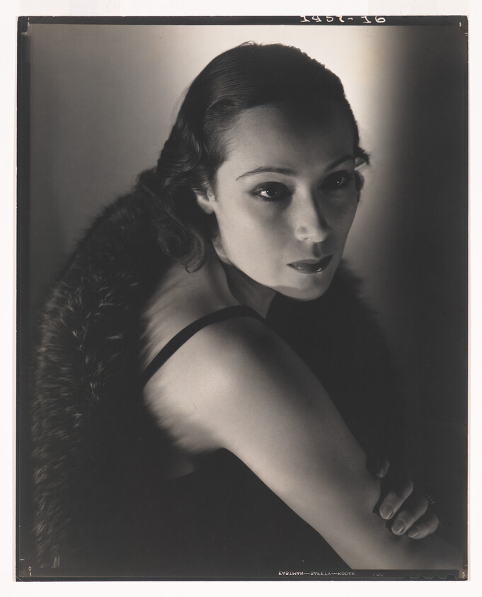 A woman in dramatic side lighting wears a fur stole and stares thoughtfully to the right.