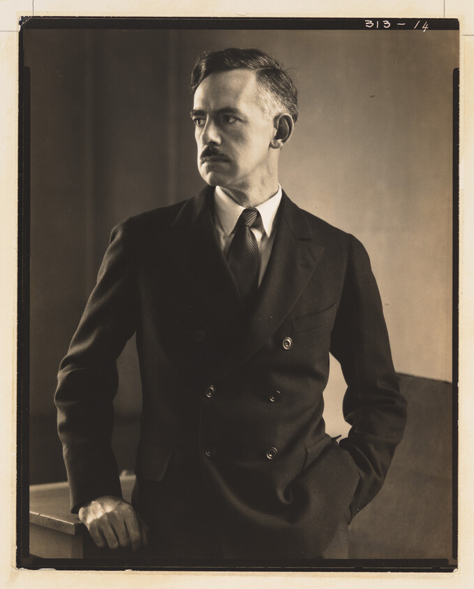A man in a double-breasted suit stands with one hand in his pocket, looking to the left.