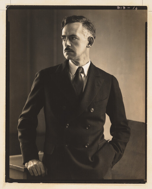 A man in a double-breasted suit stands with one hand in his pocket, looking to the left.