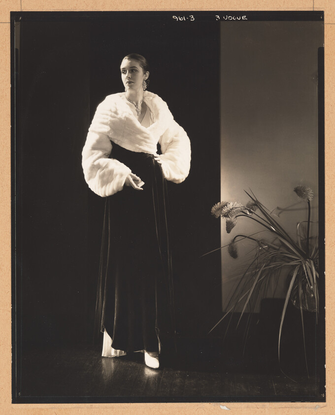 A woman in a white fur wrap and long dark gown stands posing beside a potted plant.