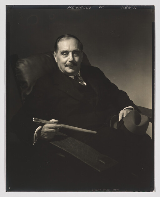 Older man in a suit sits in an armchair holding a cane and a hat.