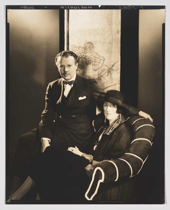 A well-dressed man stands beside a seated woman in a hat, both posing formally.