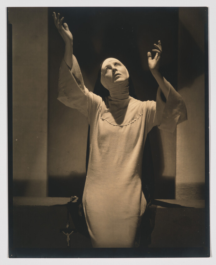 A woman in a nun's habit raises her arms and looks upward in a dramatic pose.