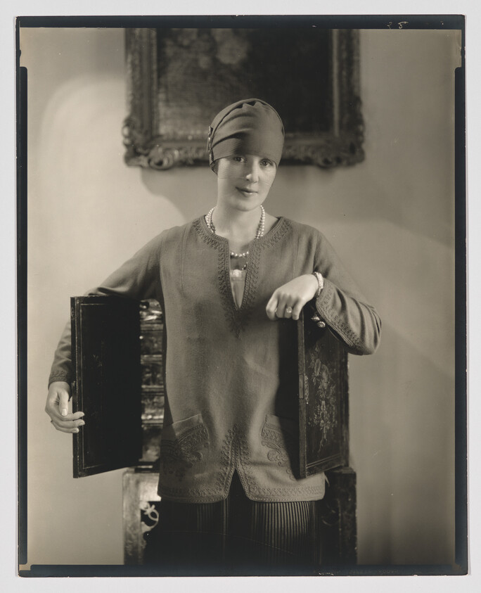 Woman in 1920s cloche hat holding open two decorative cabinet doors.