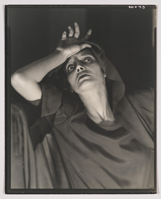 A black and white photograph of a woman with a dramatic expression, looking upwards. She is wearing a hooded garment and has her hand dramatically placed on her forehead. The lighting casts deep shadows, adding to the intensity of the scene.