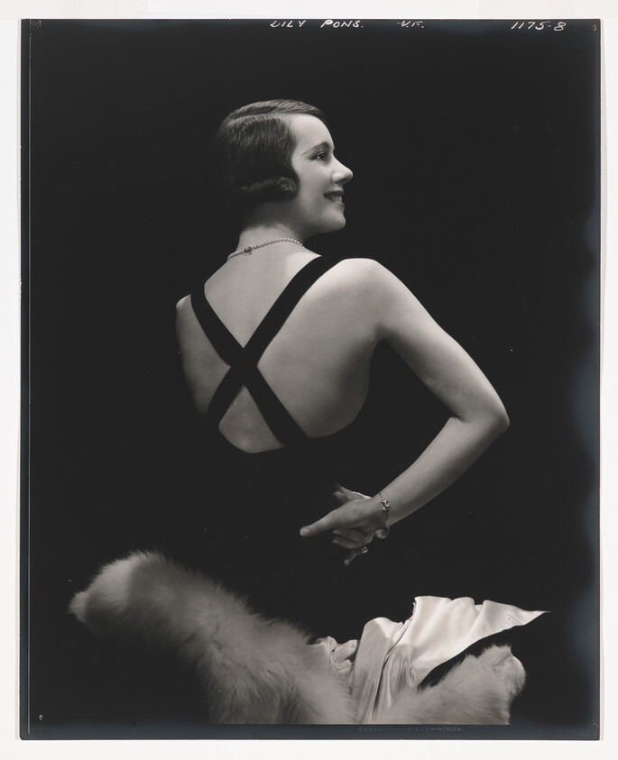 Woman in a backless dress looks over her shoulder, hands resting at her lower back.