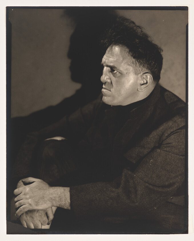 A serious man sits with hands clasped, staring sharply to the right, casting a dark shadow.
