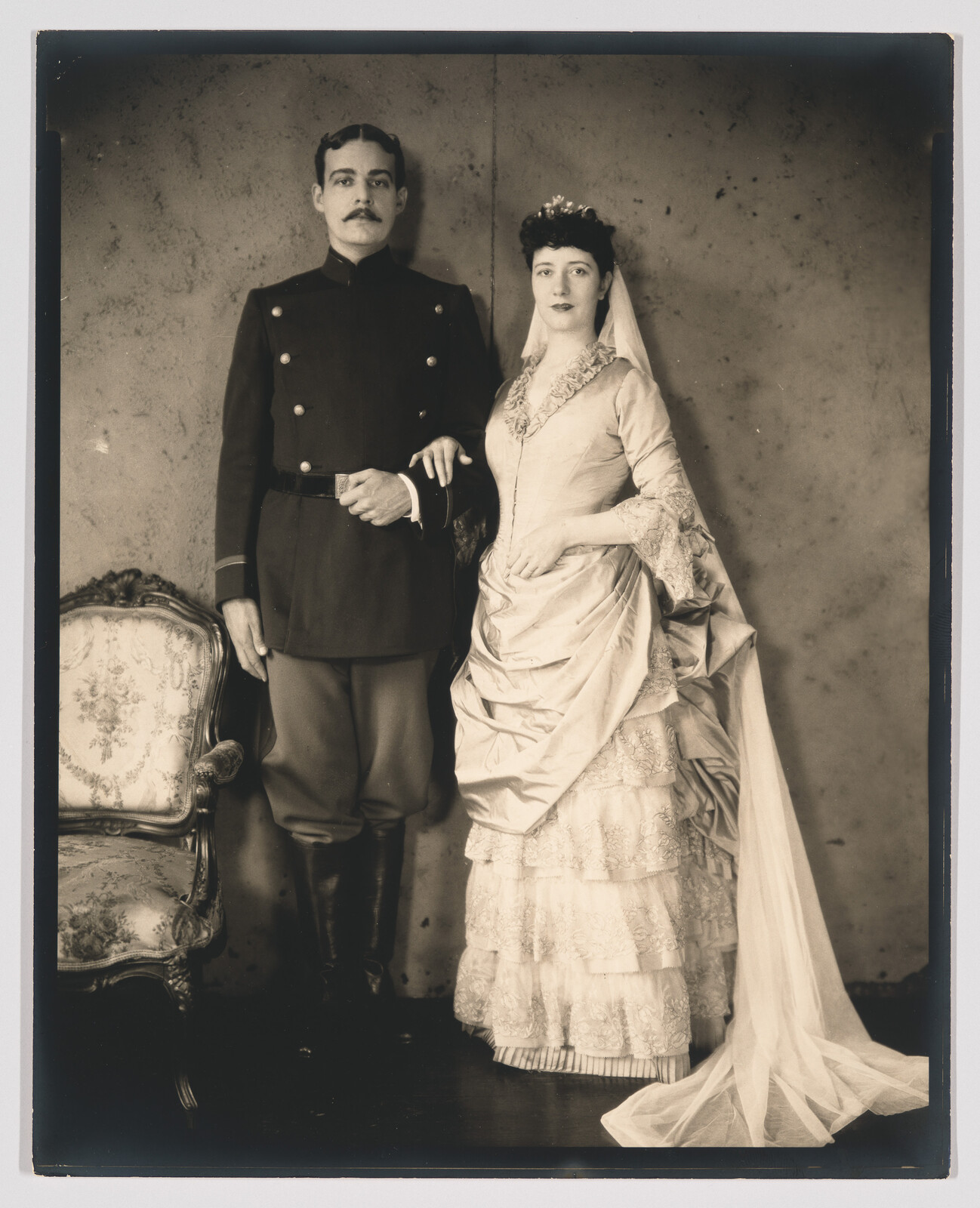A groom in a military uniform stands arm-in-arm with a bride in a lace wedding gown.