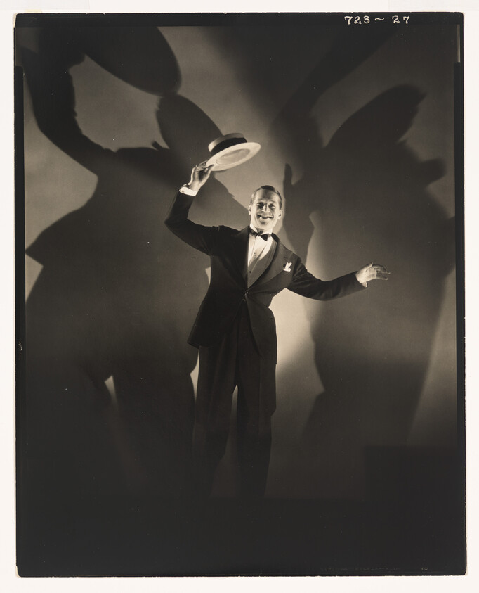 A smiling man in a tuxedo tips his hat while dramatic shadows loom behind him.