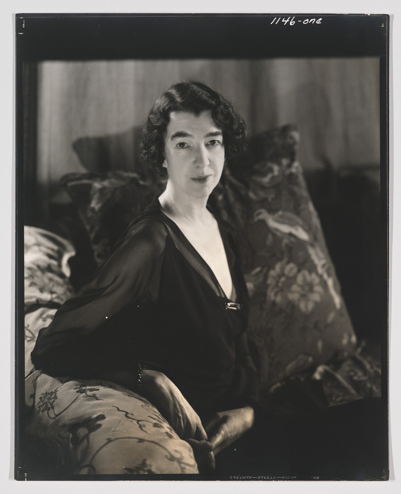 A woman in a dark dress sits on a patterned sofa, hands folded, looking toward the camera.