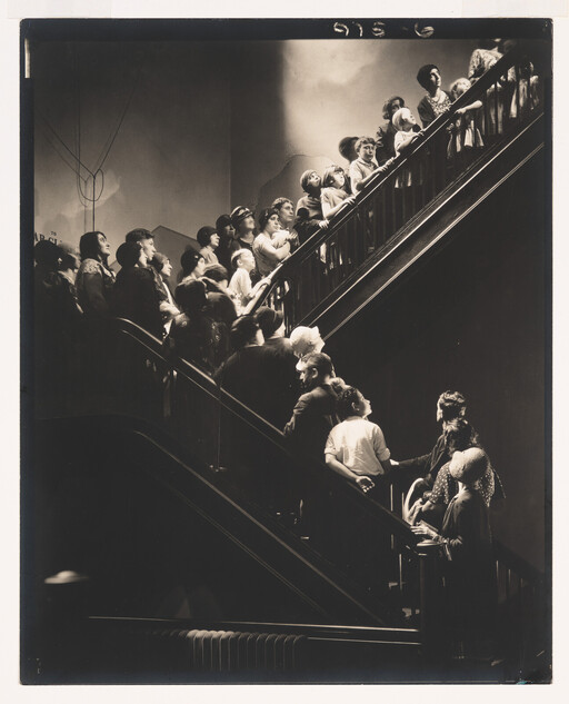A crowd of people stand on intersecting staircases, all gazing upward toward a light source.