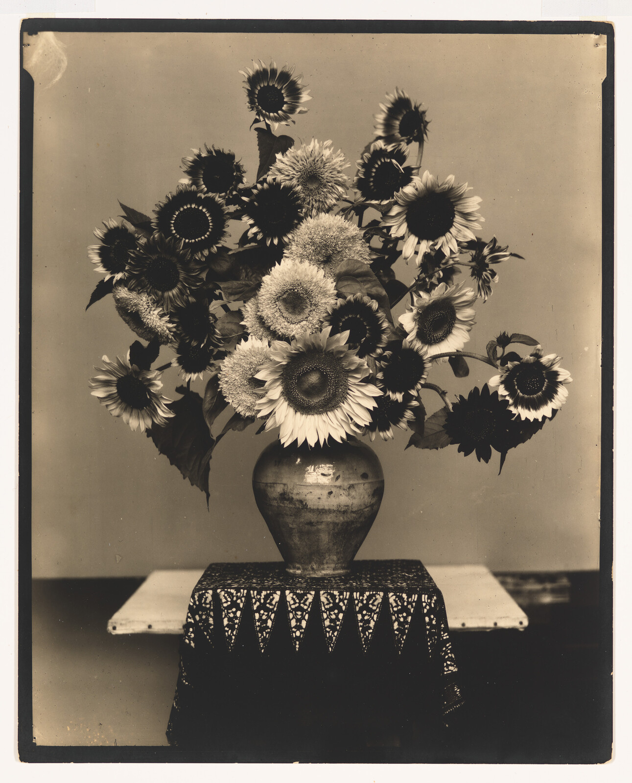 A sepia-toned photograph featuring a bouquet of sunflowers and daisies arranged in a rustic vase, which is placed on a table with a decorative lace tablecloth. The image has a vintage appearance with soft lighting that enhances the textures of the flowers and vase.