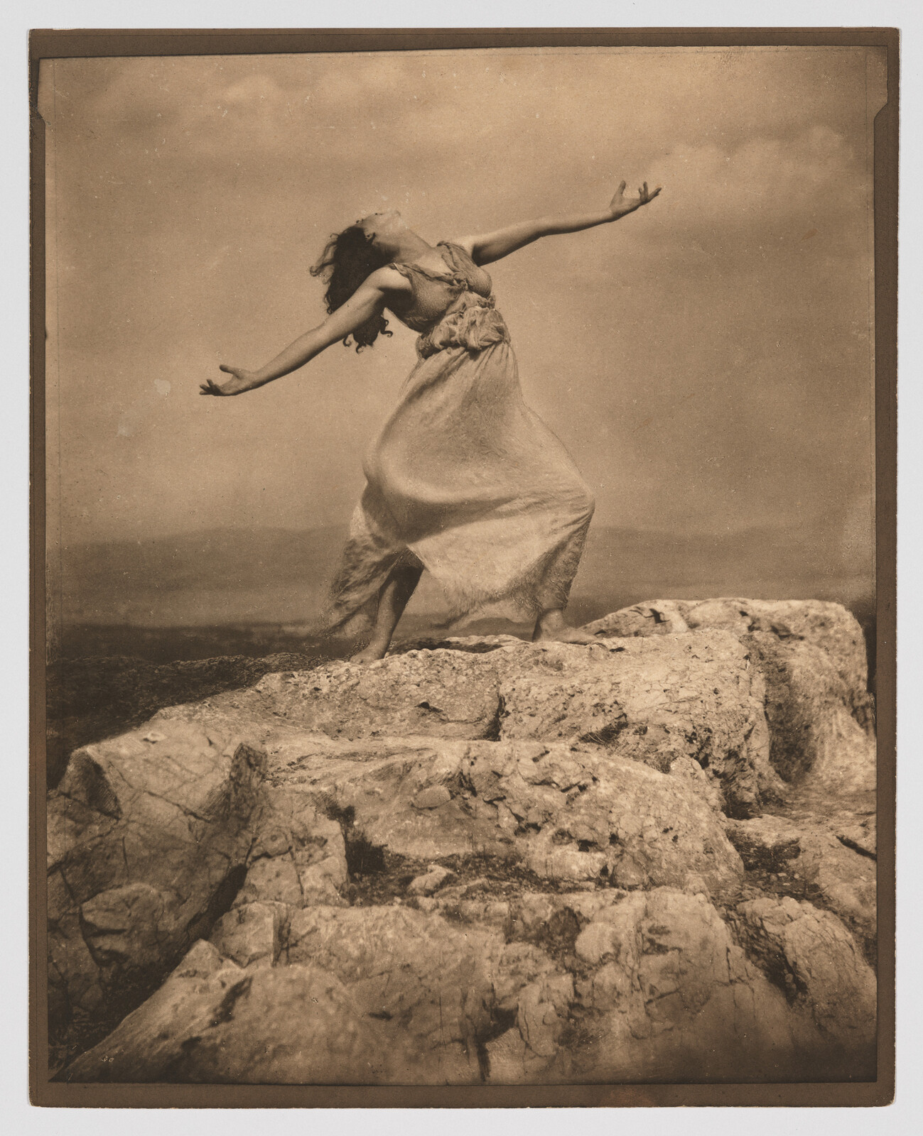 A woman in a flowing dress stands on rocky cliffs with arms outstretched toward the sky.