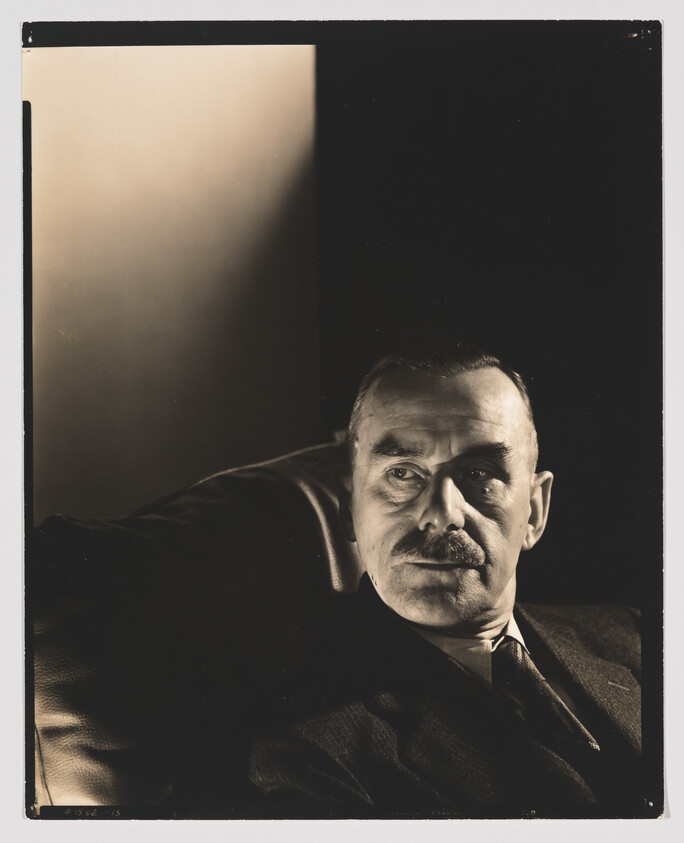 A middle-aged man with a mustache sits in shadowed light, looking thoughtfully to the side.