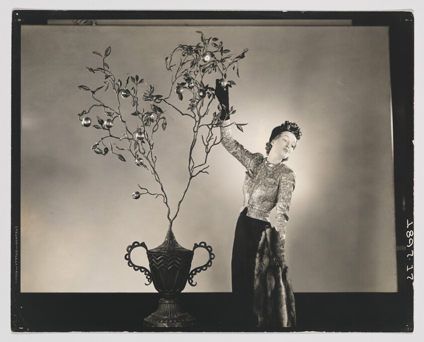 A woman in vintage dress reaches up to adjust ornaments on a tall decorative branch in a vase.