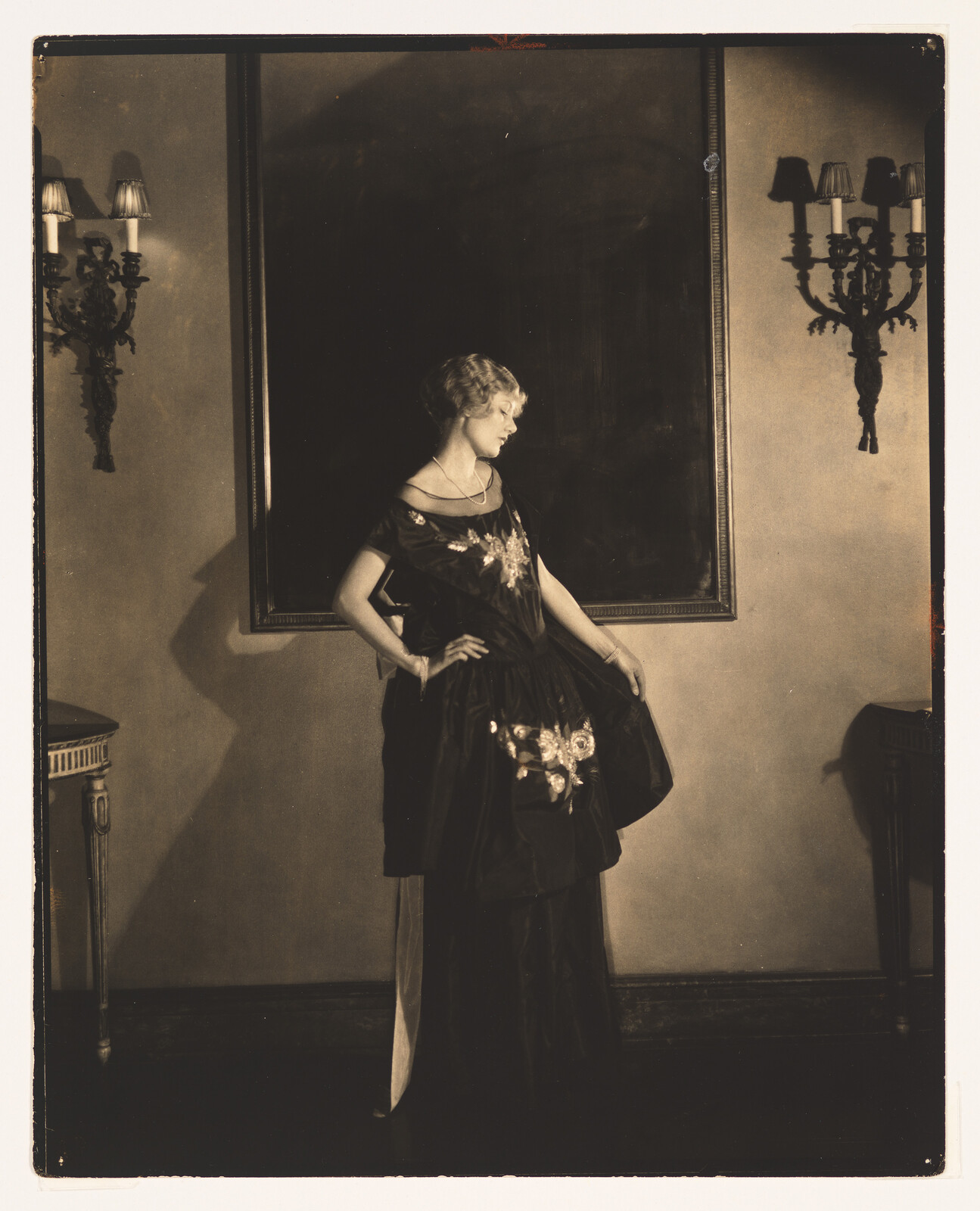 Woman in an ornate dark dress poses with hands on hips in an elegant room.