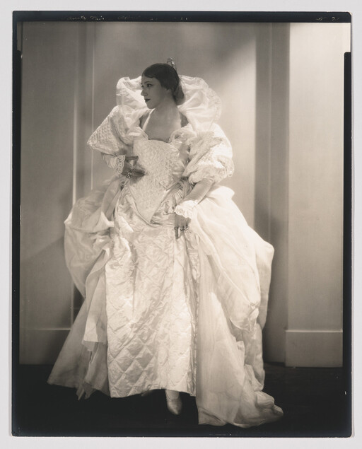 Woman wearing an elaborate voluminous white gown and gloves poses while looking to her left.