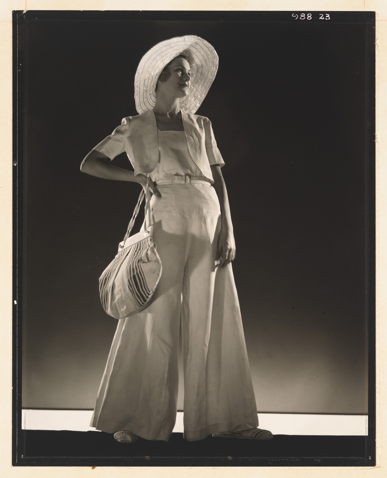 Young woman stands with hand on hip wearing a wide-brim hat, wide trousers, and striped bag.