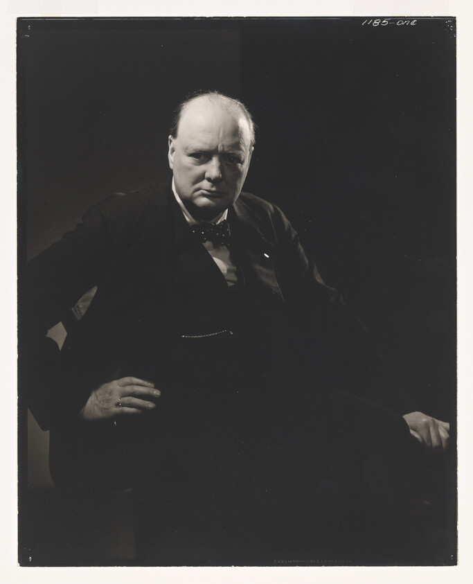 Older balding man in a dark suit and bow tie sits with hands on hips, looking serious.