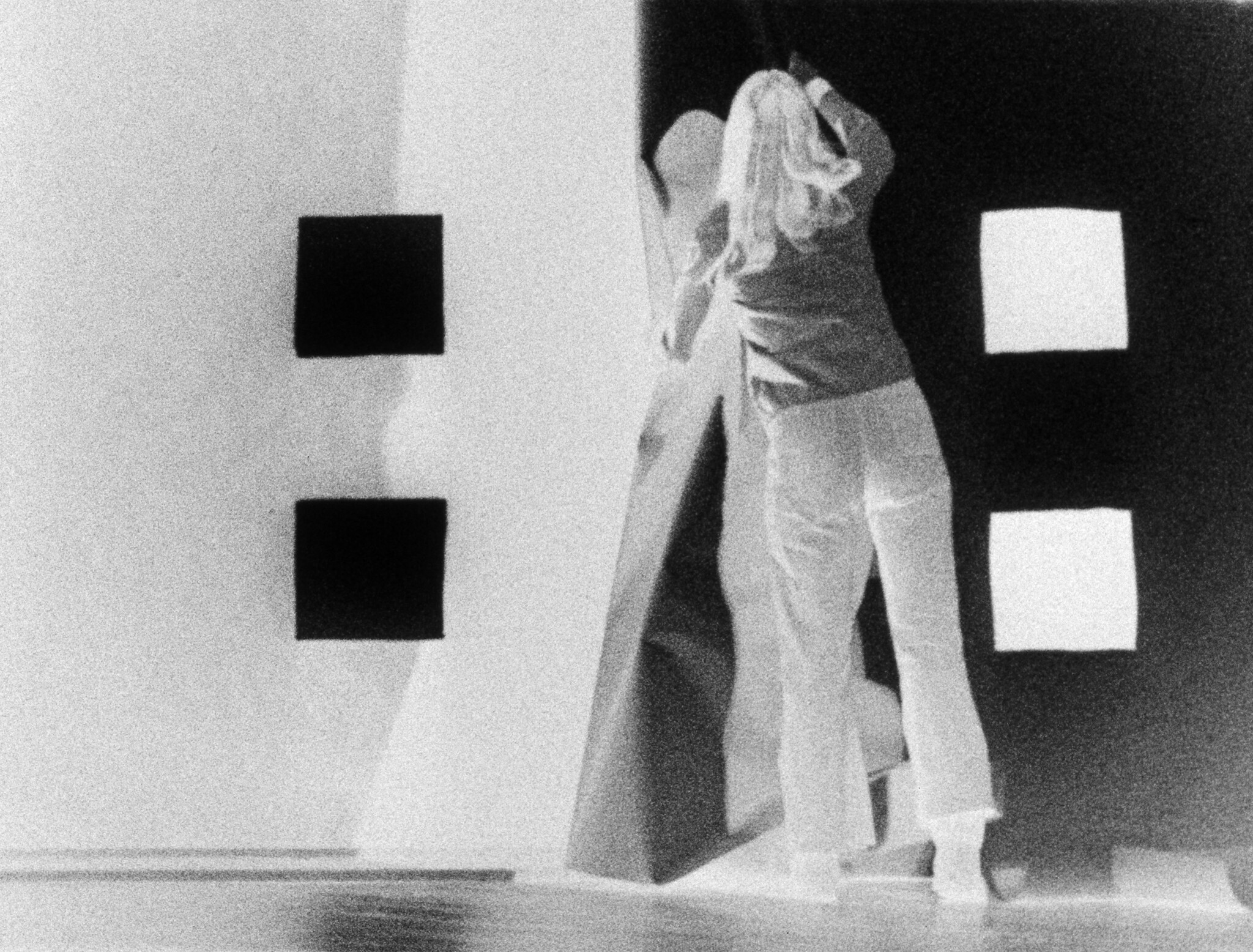 Person with long hair stands facing a wall with square shapes, reaching up with one arm.