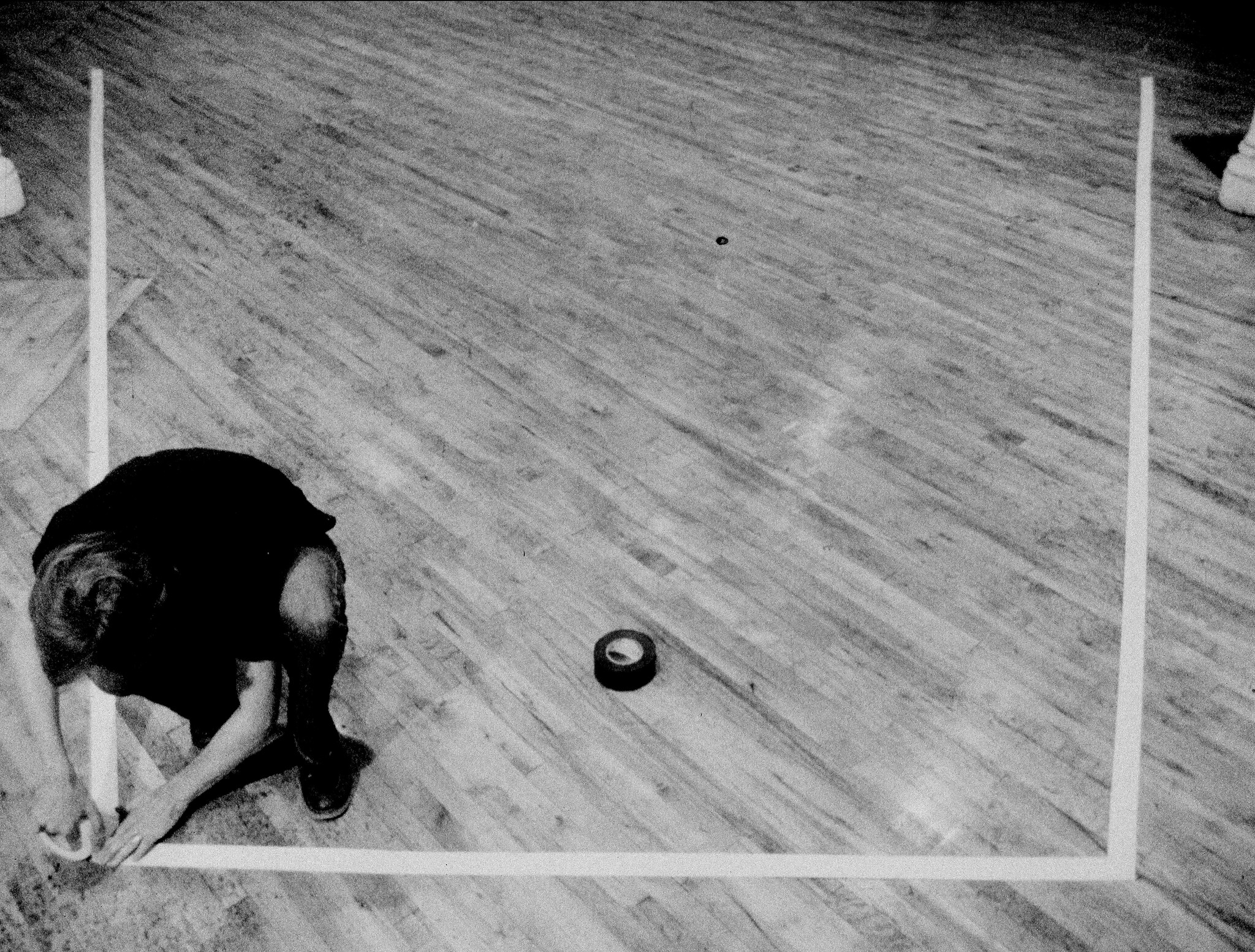Person crouching on a wooden floor, placing tape to form a large square outline with a roll nearby.